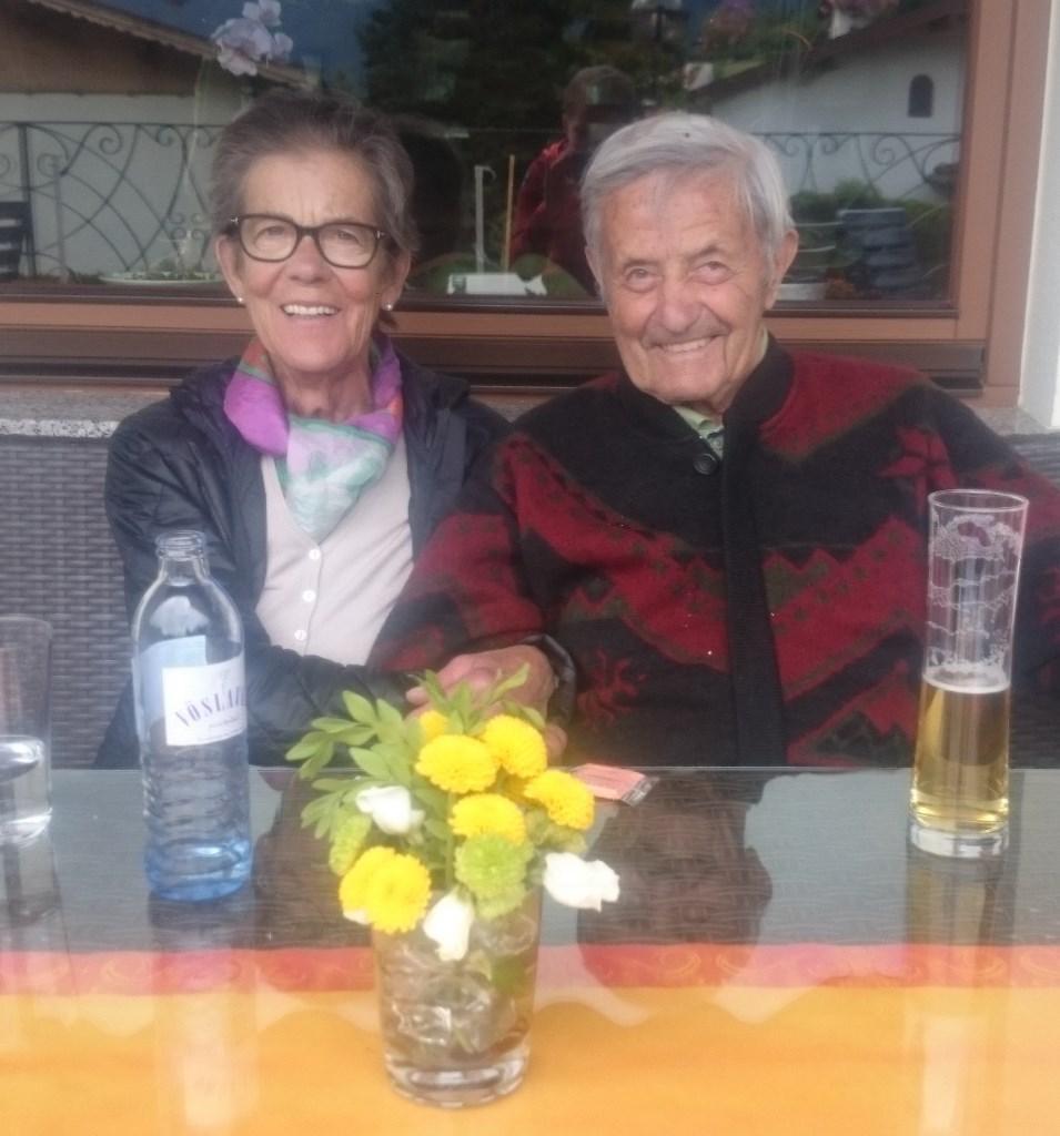 Roswitha und Sepp sitzen hinter einem Tisch. Am Tisch vor ihnen steht eine Vase mit gelben Blumen. Sepp trinkt ein Bier. Roswitha ein Mineralwasser.
