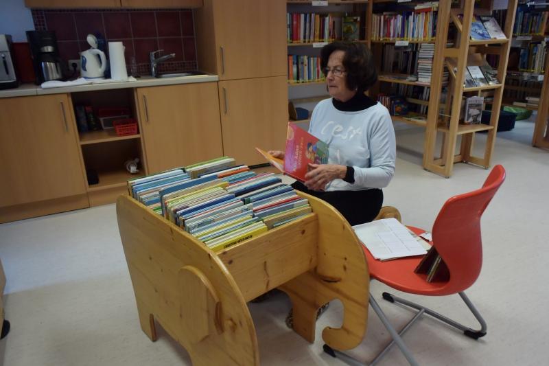 Lesepatin Inge sitzt hinter einem Büchertrog und hält ein Buch in der Hand