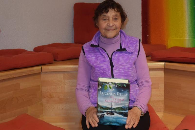 Mitarbeitertin Anneliese sitzt auf einer Eckbank und hält ein Buch auf den Oberschenkeln